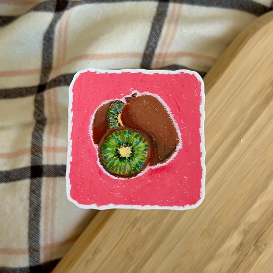 Sticker of a sliced kiwi on a pink background, placed on a wooden surface with plaid fabric underneath.