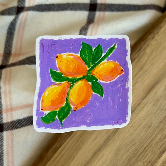 Square tile with a painting of lemons on a purple background, placed on a checkered fabric surface.