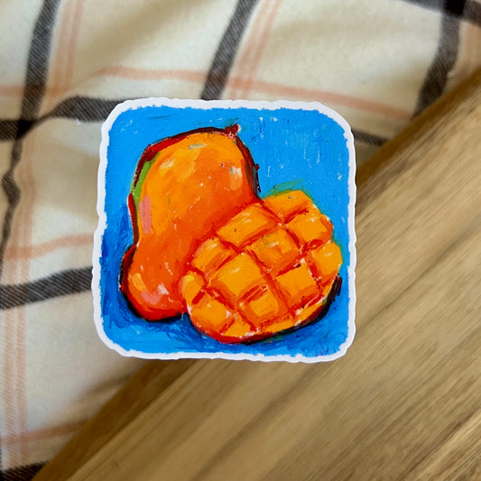 Small painting of a mango on a blue background, placed on a wooden surface with a plaid fabric background.