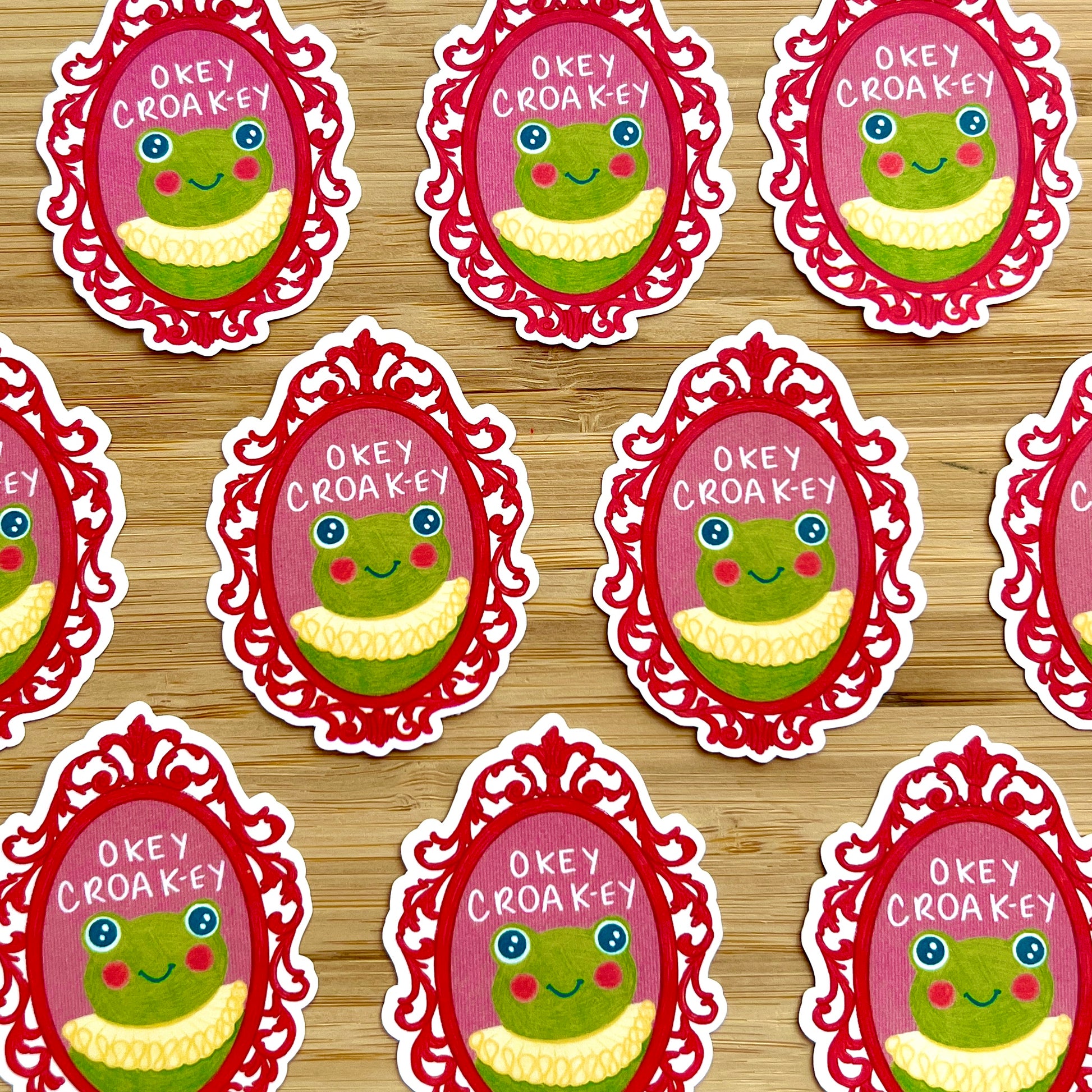 Repeating pattern of cartoon frog character stickers with 'Okey Croakey' text on a wooden surface.