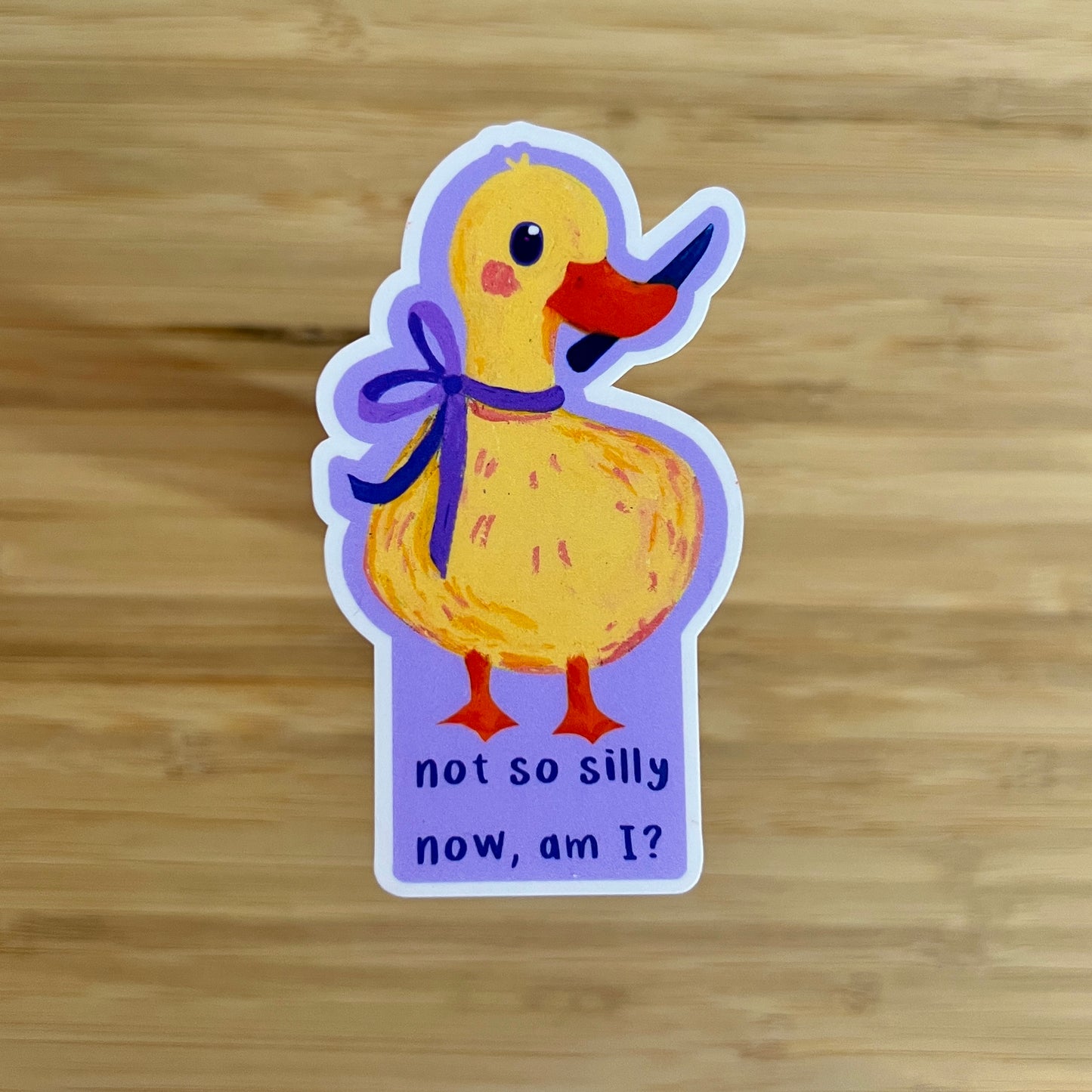 Sticker of a yellow duck with a purple bow and text on a wooden surface