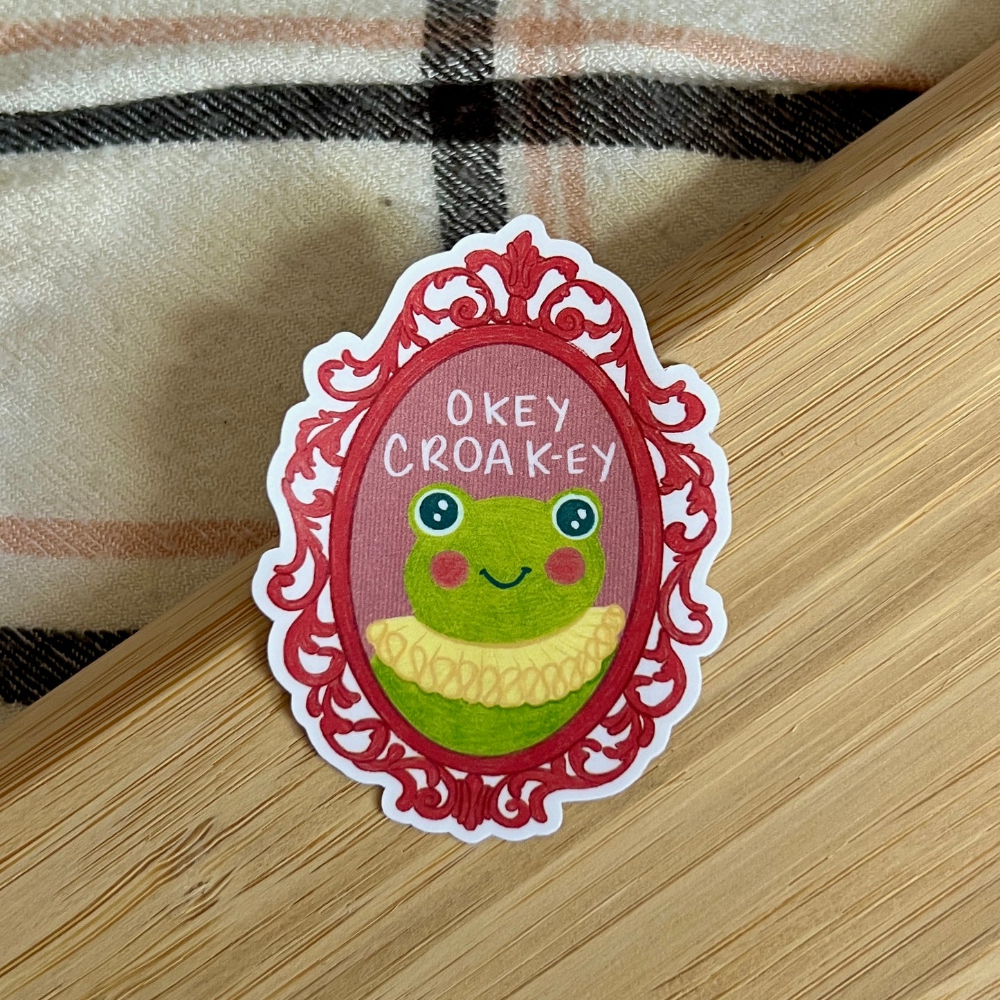 Sticker with a frog design and text 'Okey Croak-ey' on a wooden surface.
