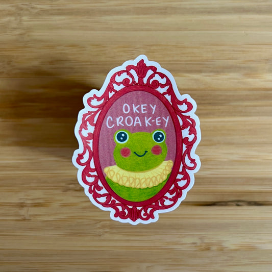 Sticker with a frog design and text 'Okey Croak-ey' on a wooden surface