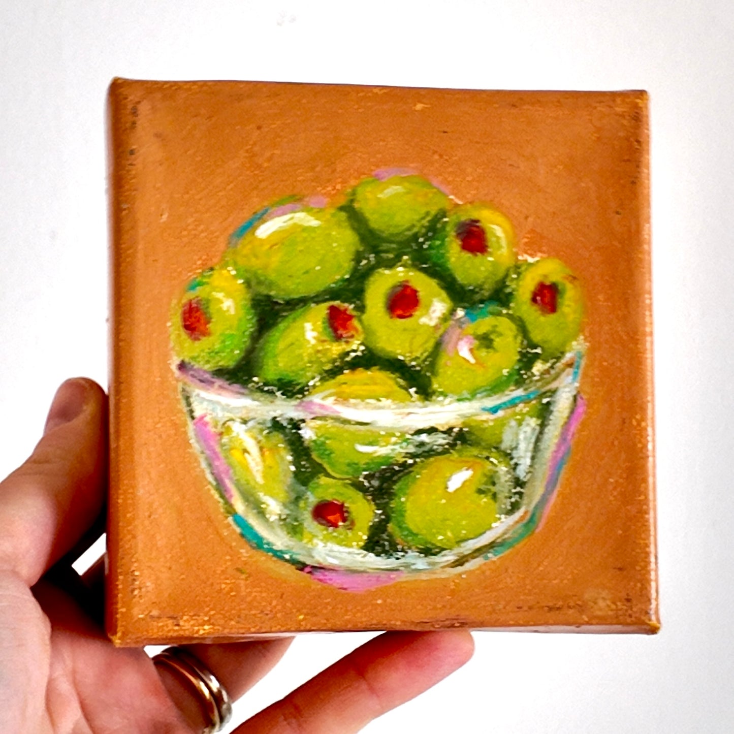 Small painting of green olives in a glass jar held by a hand.