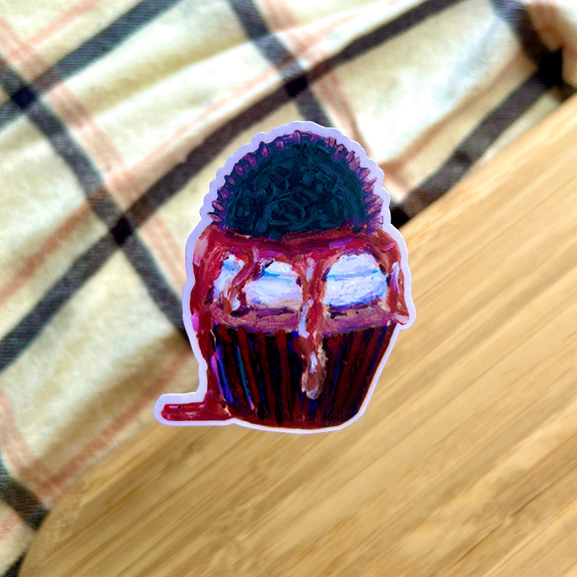Sticker of a cupcake with purple frosting and chocolate drizzle on a checkered fabric background.