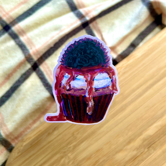 Sticker of a cupcake with purple frosting and chocolate drizzle on a checkered fabric background.