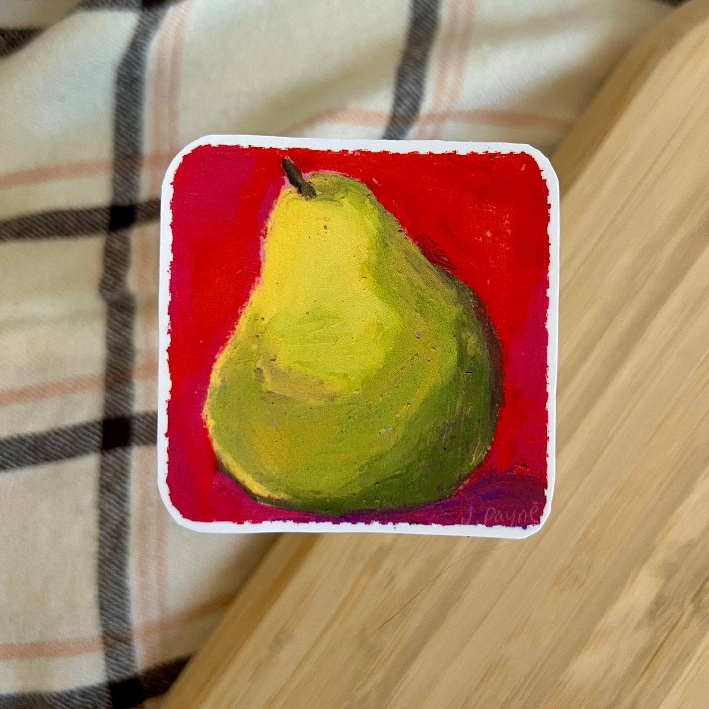 Painting of a green pear on a red background, placed on a wooden surface with a plaid fabric.
