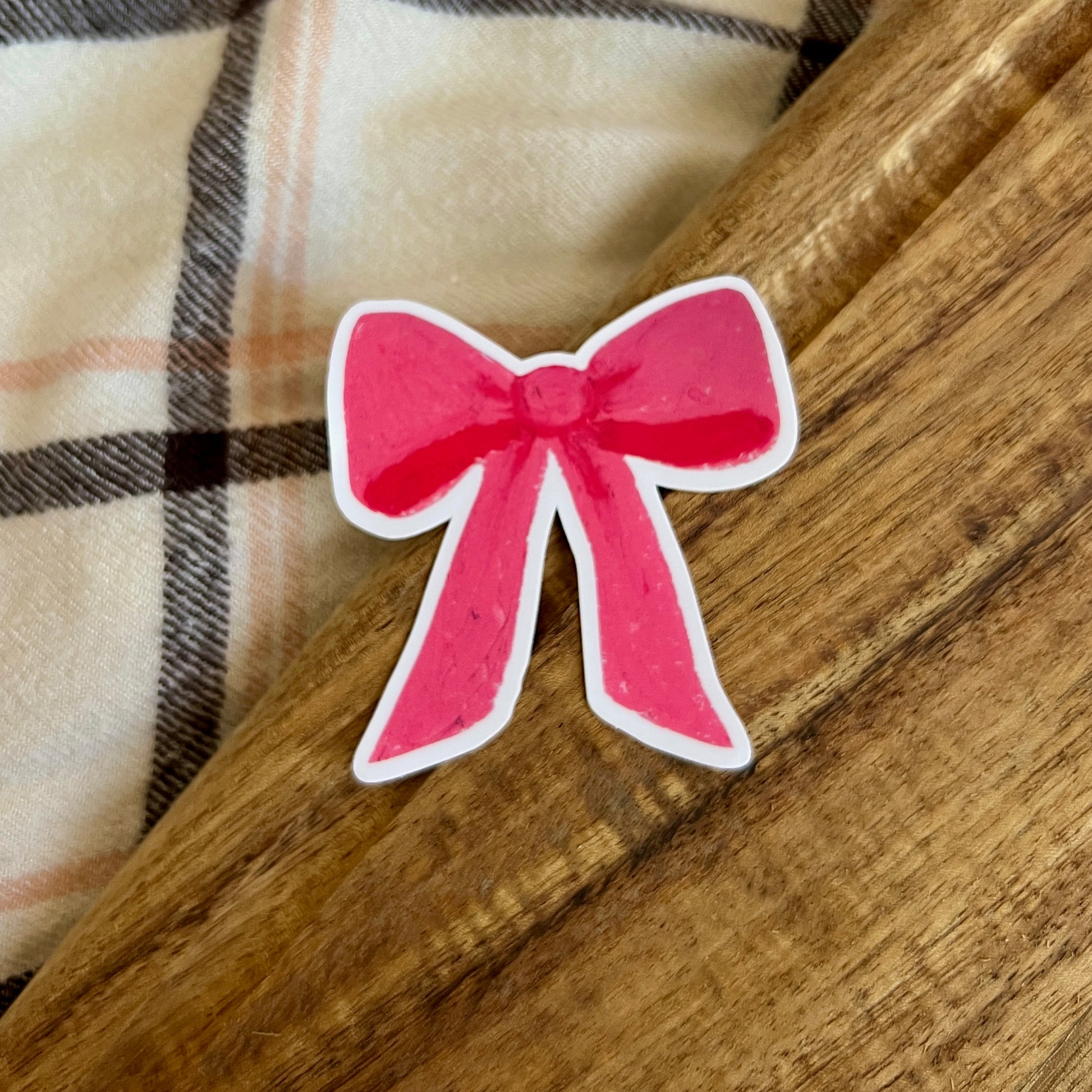 Pink bow sticker on a wooden surface with plaid fabric in the background
