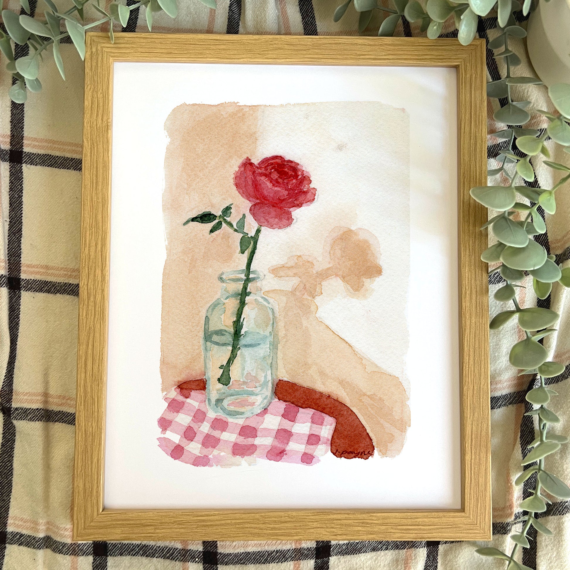 Framed artwork of a red rose in a glass jar on a checkered cloth with a wooden frame.