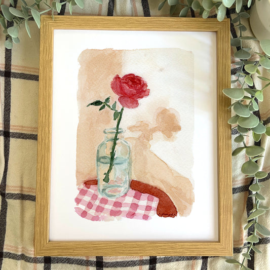 Framed artwork of a red rose in a glass jar on a checkered cloth with a wooden frame.
