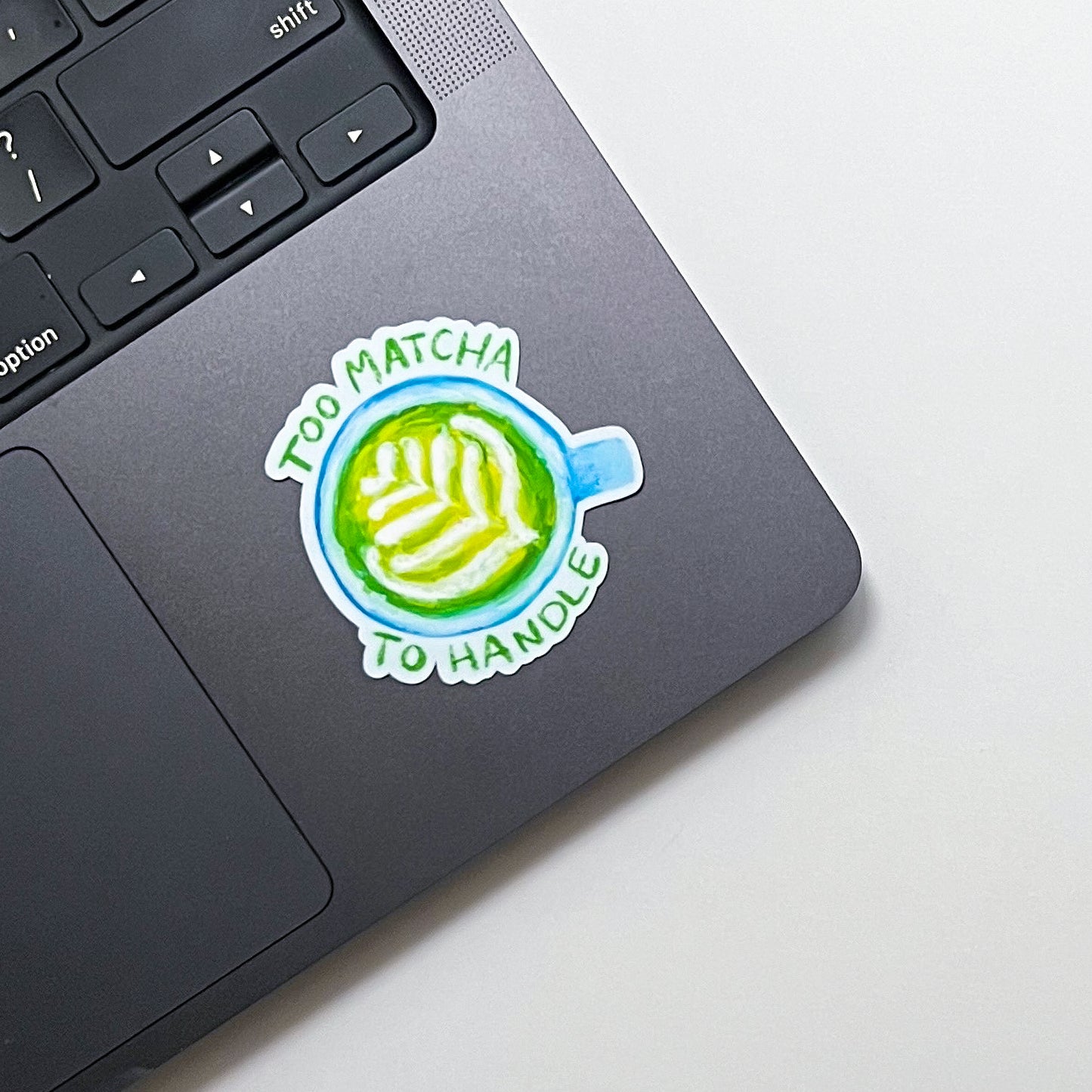 Laptop with a sticker that reads 'Too Matcha To Handle' on a white surface