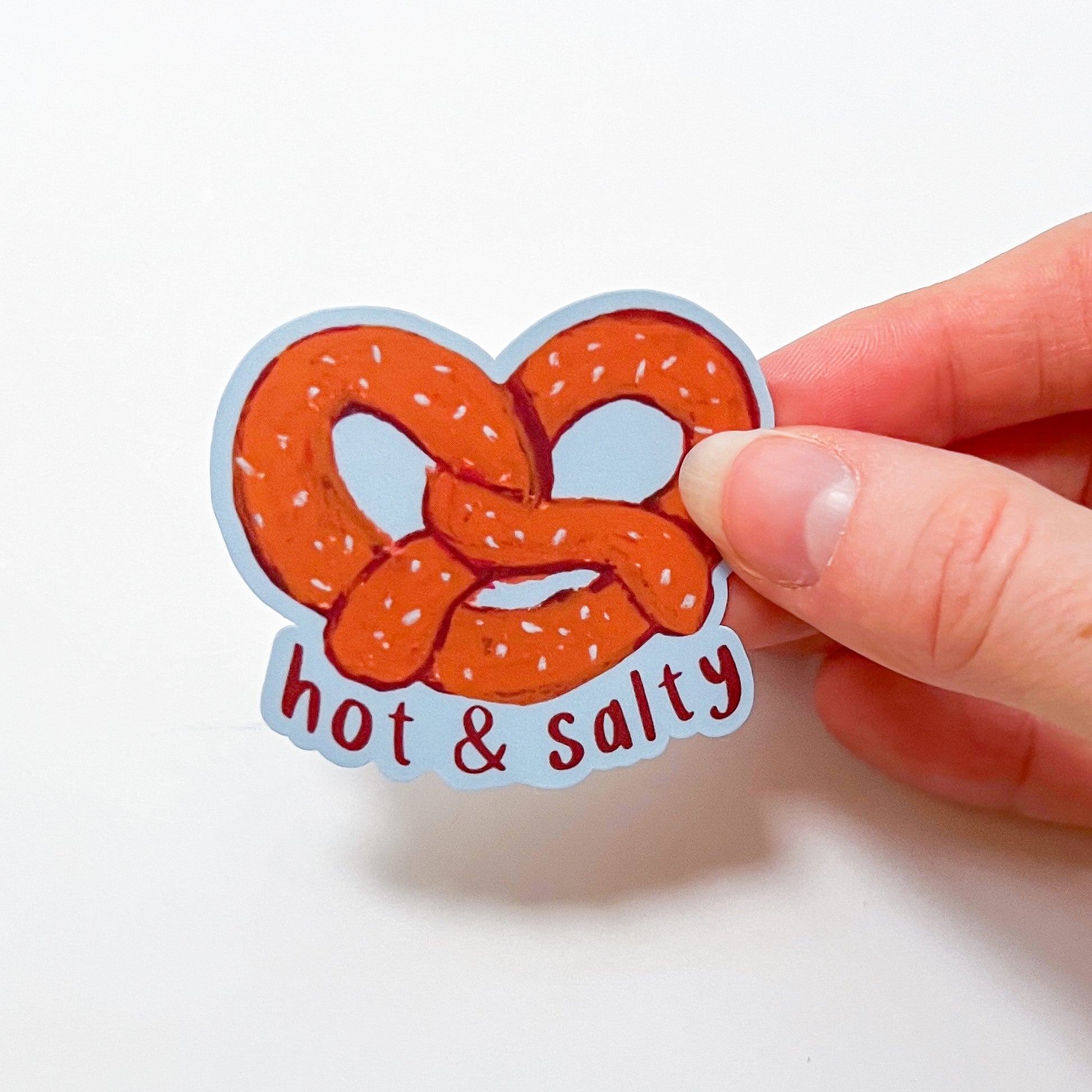 Hand holding a sticker of a pretzel with 'hot & salty' text on a white background