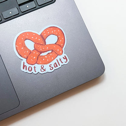 Laptop with a sticker of a pretzel and 'hot & salty' text on a white surface