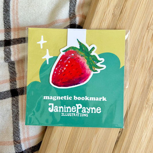 Magnetic bookmark with strawberry illustration and Janine Payne branding on a wooden surface.
