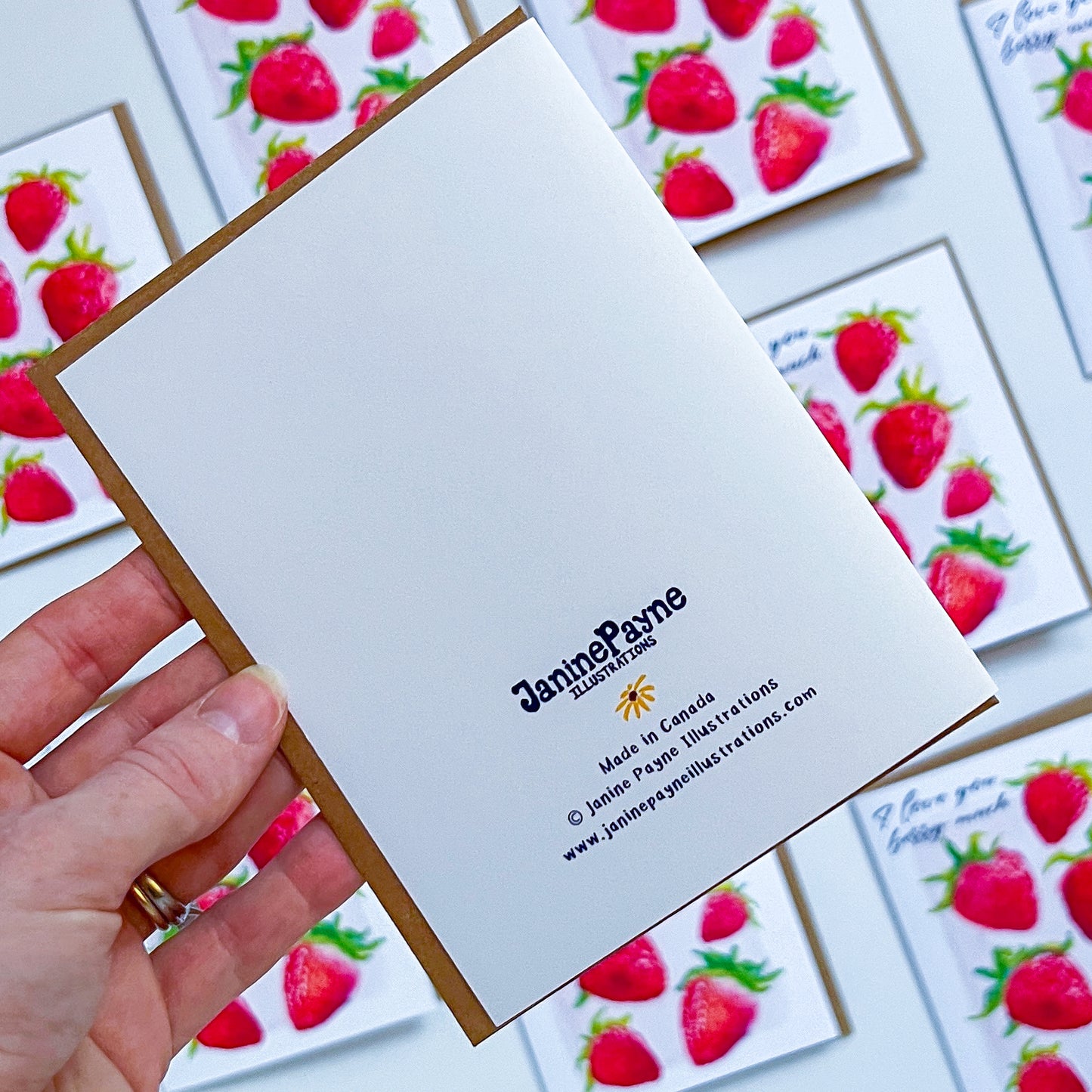 Hand holding a card with 'Janine Payne Illustrations' branding, surrounded by strawberry-themed cards.