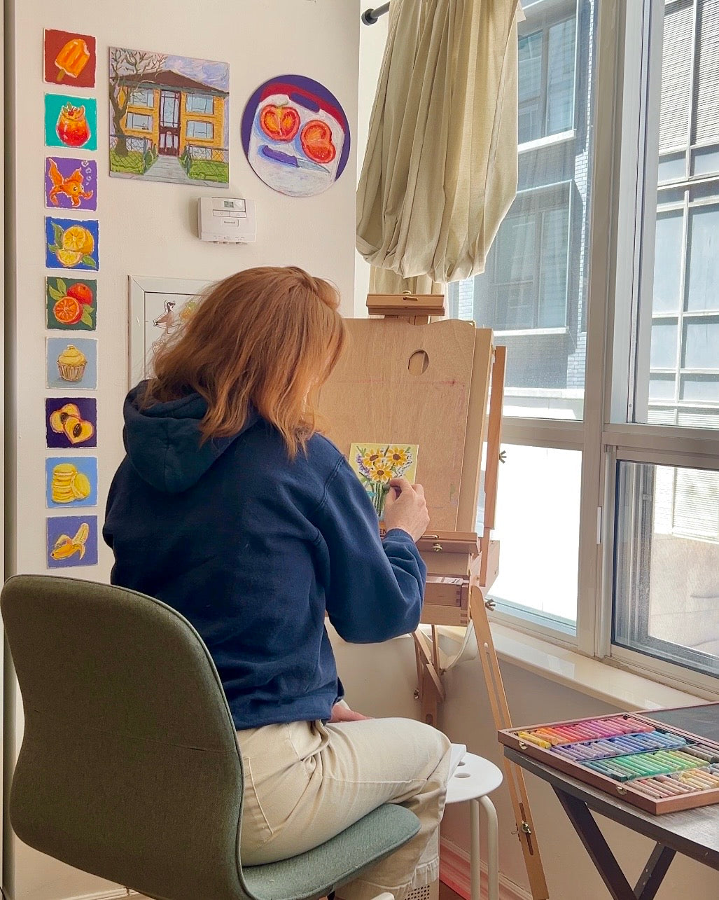 Person painting on an easel in a room with colorful paintings on the wall.