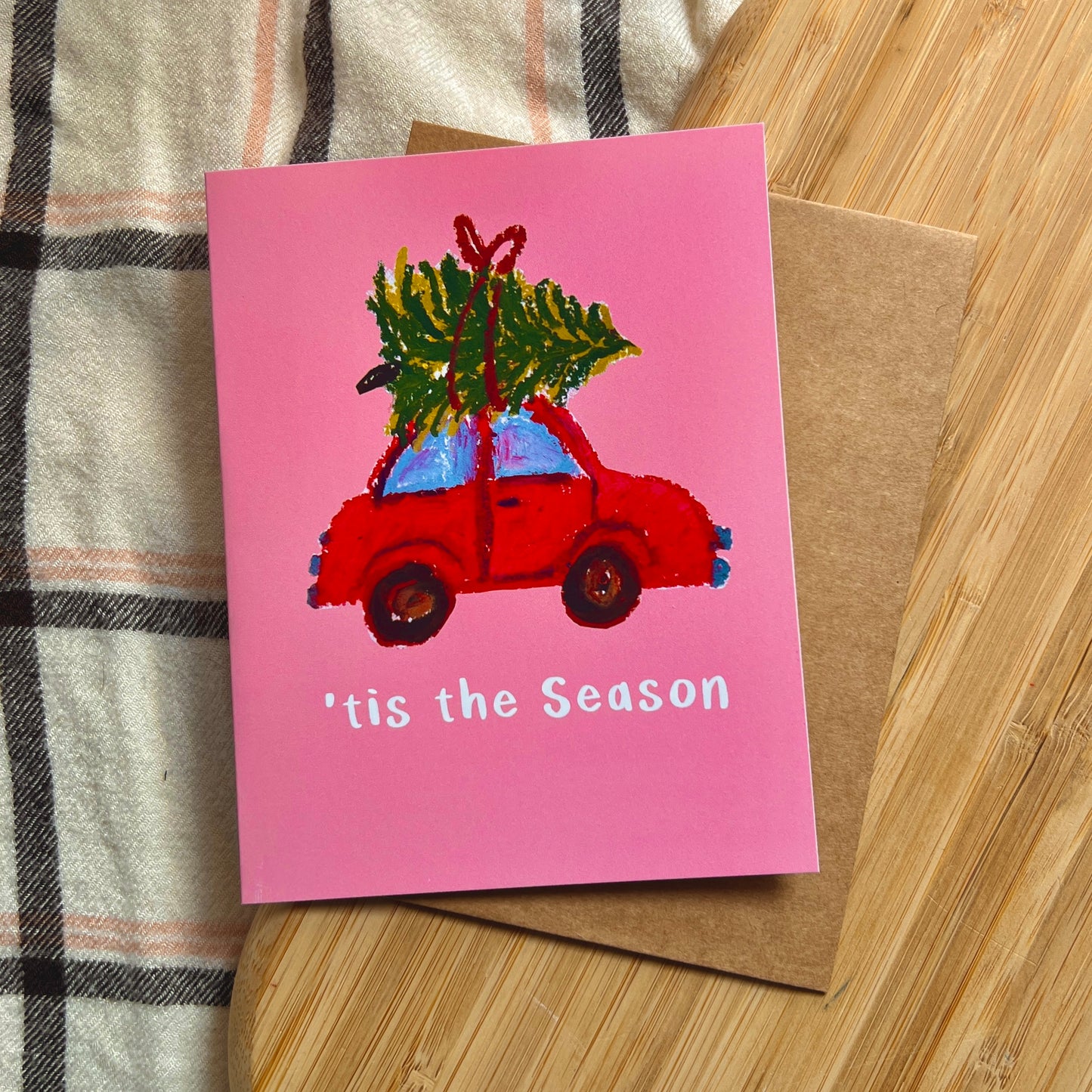 Pink Christmas card with a red car carrying a tree on a wooden surface.