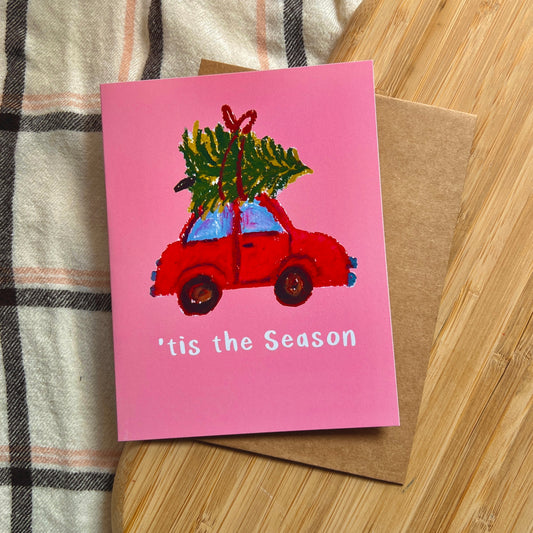 Pink Christmas card with a red car carrying a tree on a wooden surface.
