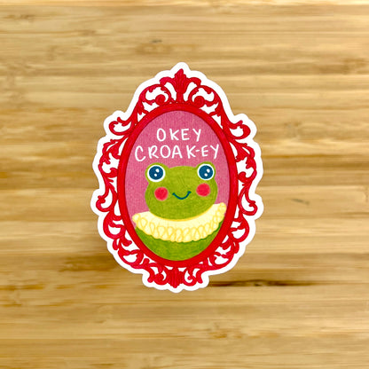 Sticker with a frog design and 'Okey Croak-ey' text on a wooden background
