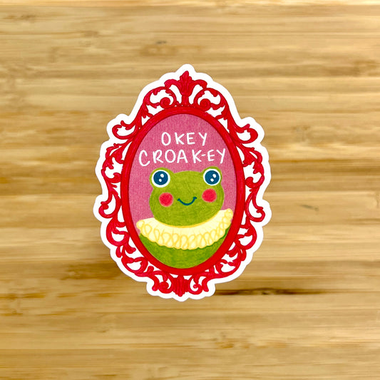Sticker with a frog design and 'Okey Croak-ey' text on a wooden background