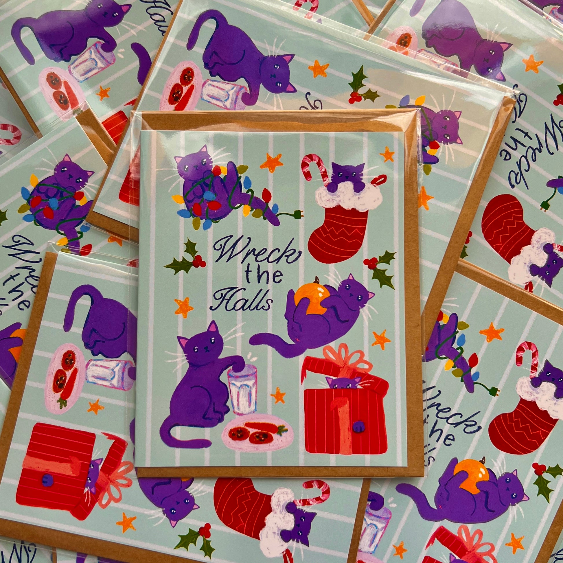 Stack of greeting cards with purple cats and Christmas-themed illustrations on a light blue background.