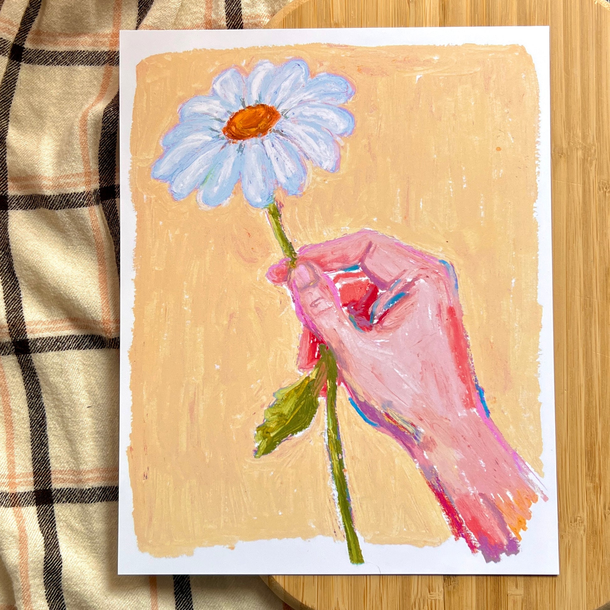 illustrated hand holding daisy with yellow background on wooden and plaid surface